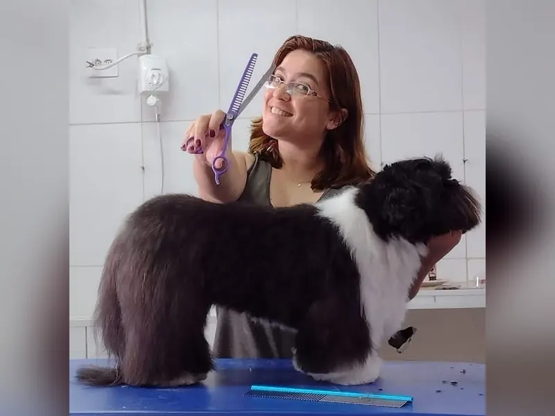 Profissional do Mogi Dog sorrindo com uma tesoura de tosa ao lado de um cão de pelo preto e branco sobre a mesa de tosa